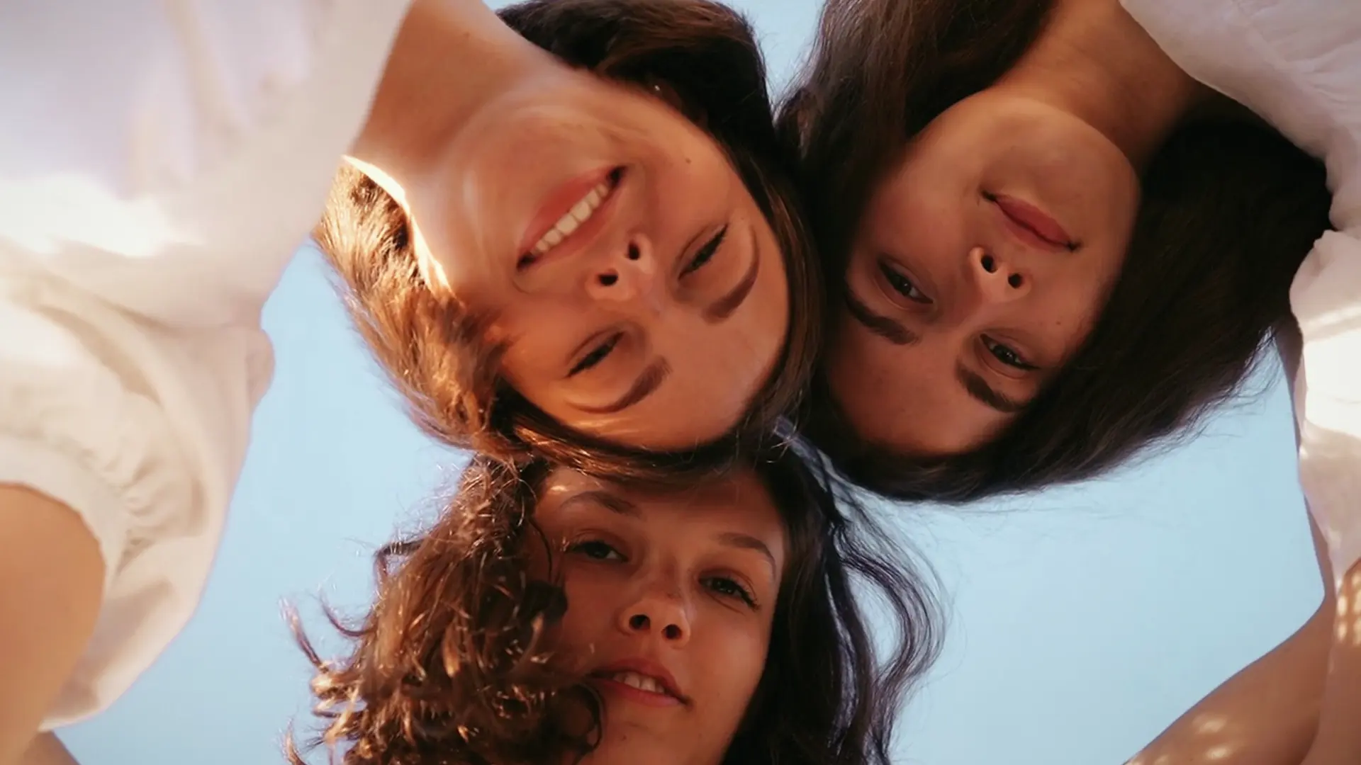 Trois jeunes filles allongées en cercle regardent vers le haut, sur fond de ciel bleu