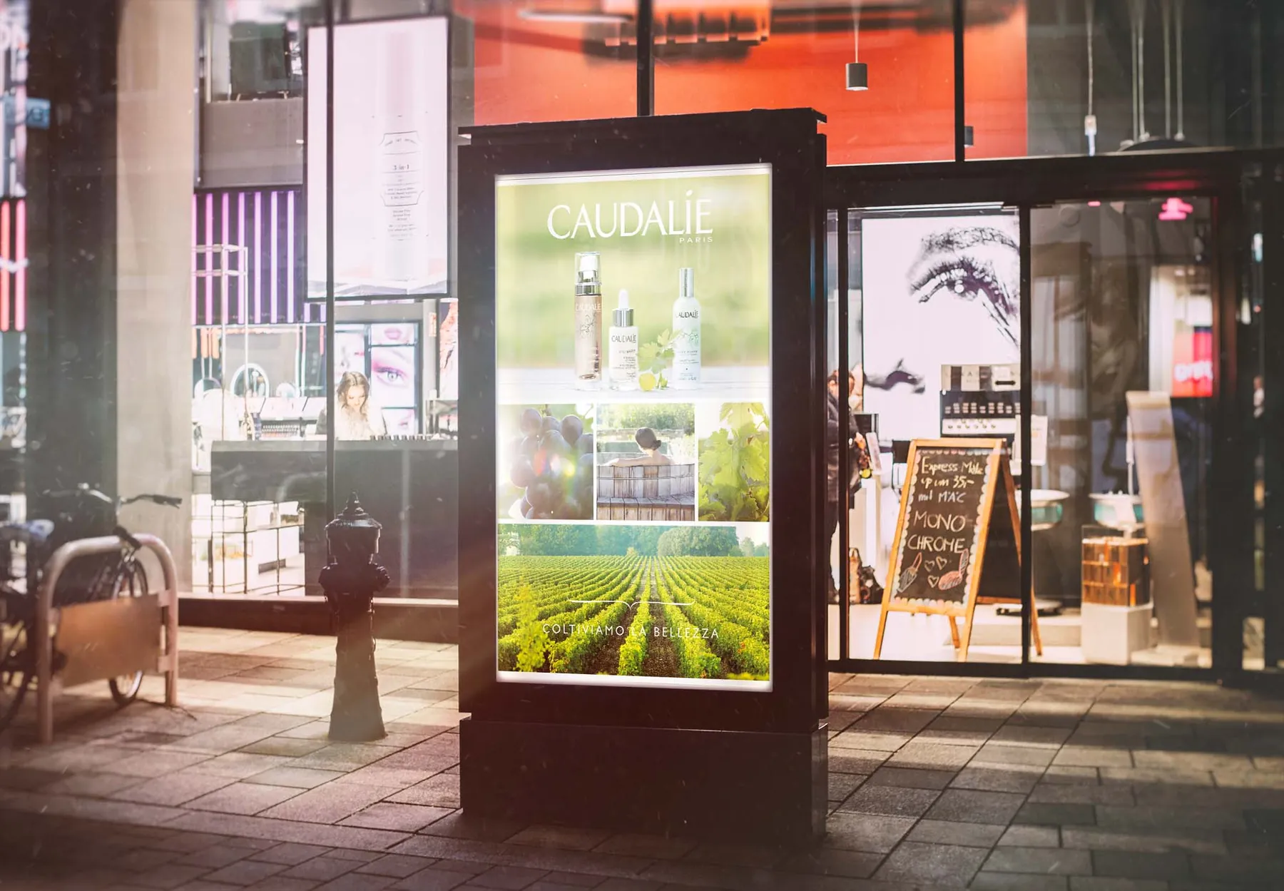 Le poster Caudalie sur un totem lumineux posé sur le trottoir, le soir, devant un magasin éclairé ; à côté une ardoise couverte d’inscriptions à la craie