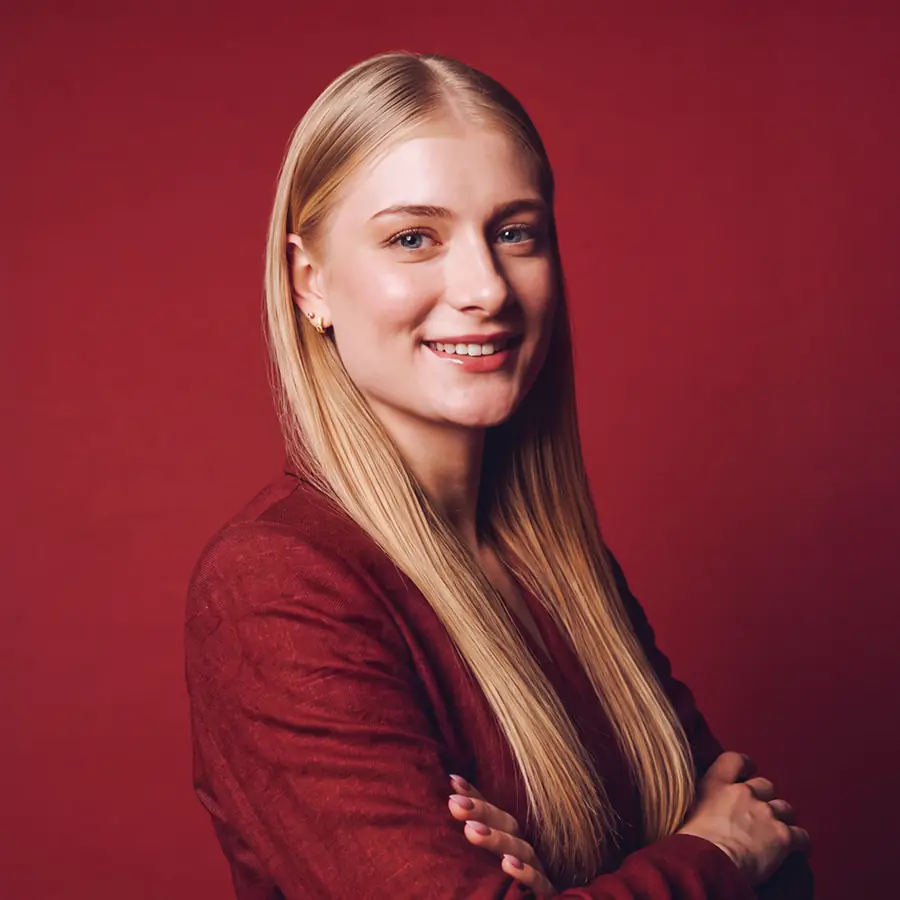 Portrait de trois quarts d’une jeune femme blonde en veste bordeaux, sur fond rouge