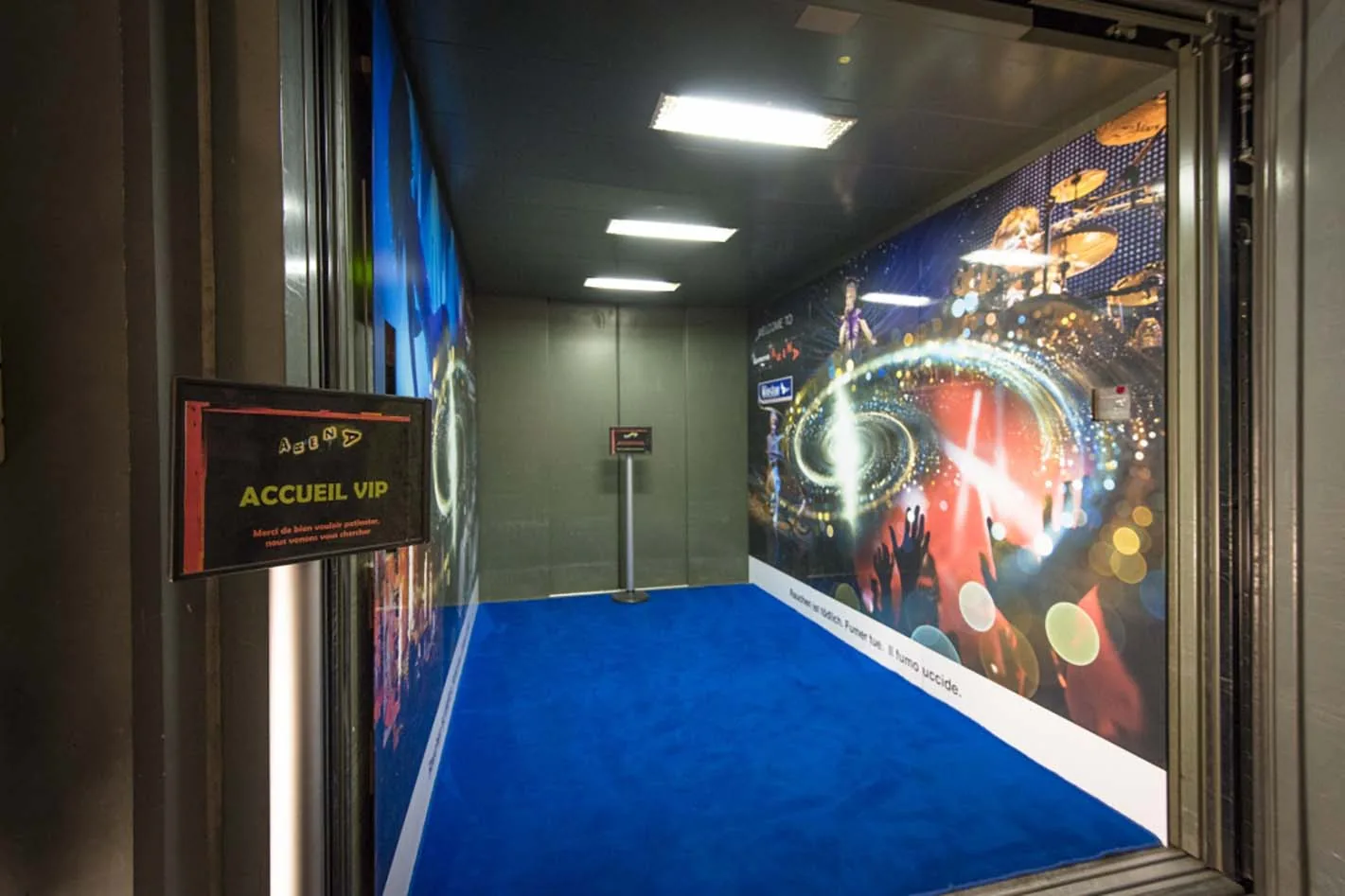 L’entrée de l’ascenseur VIP : une cabine à moquette bleue et une paroi couverte d’une image de concert avec une spirale lumineuse ; à gauche un panneau portant l’inscription Accueil VIP