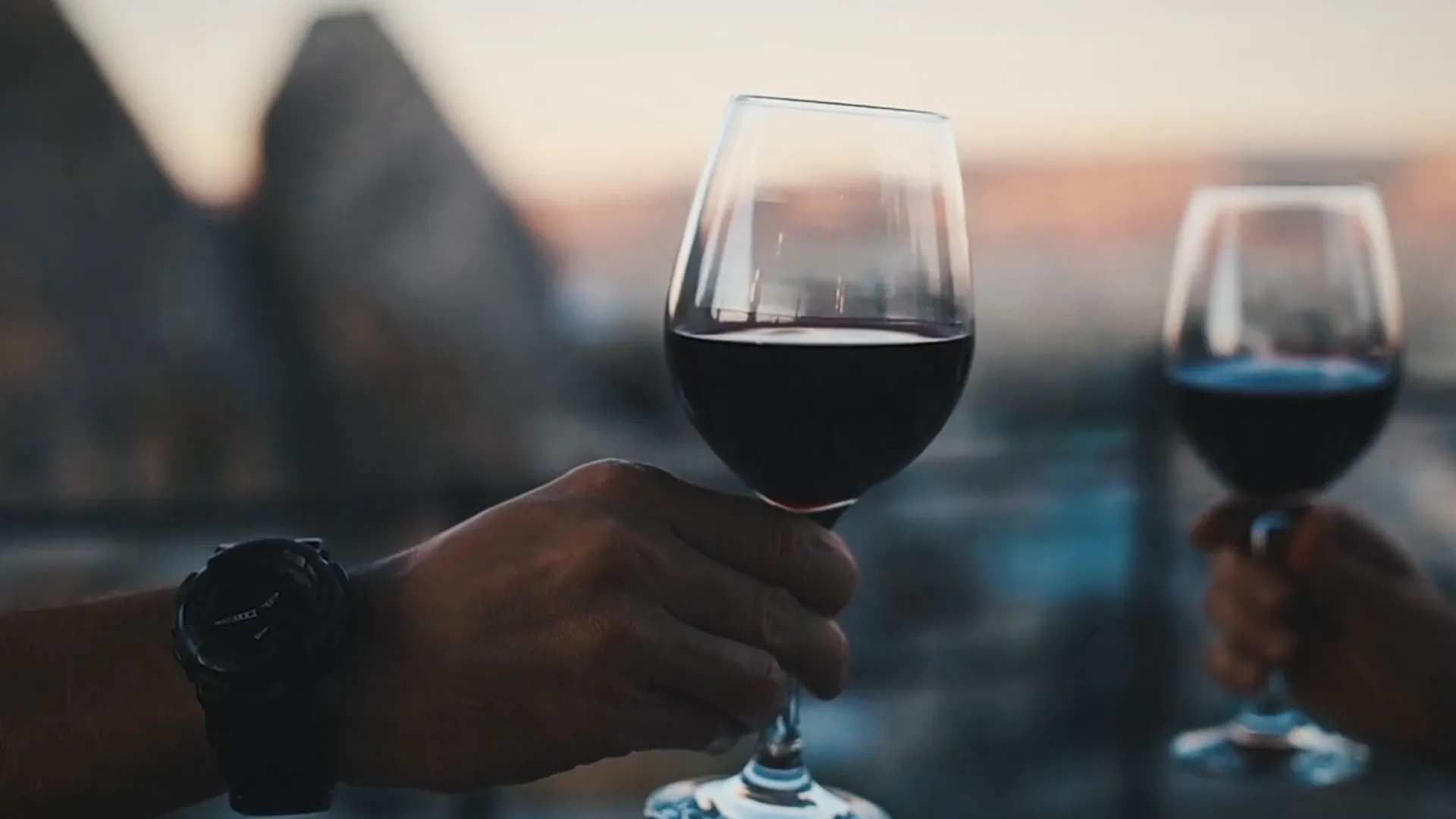 Due mani alzano due calici di vino rosso al tramonto, con lo sfondo sfocato