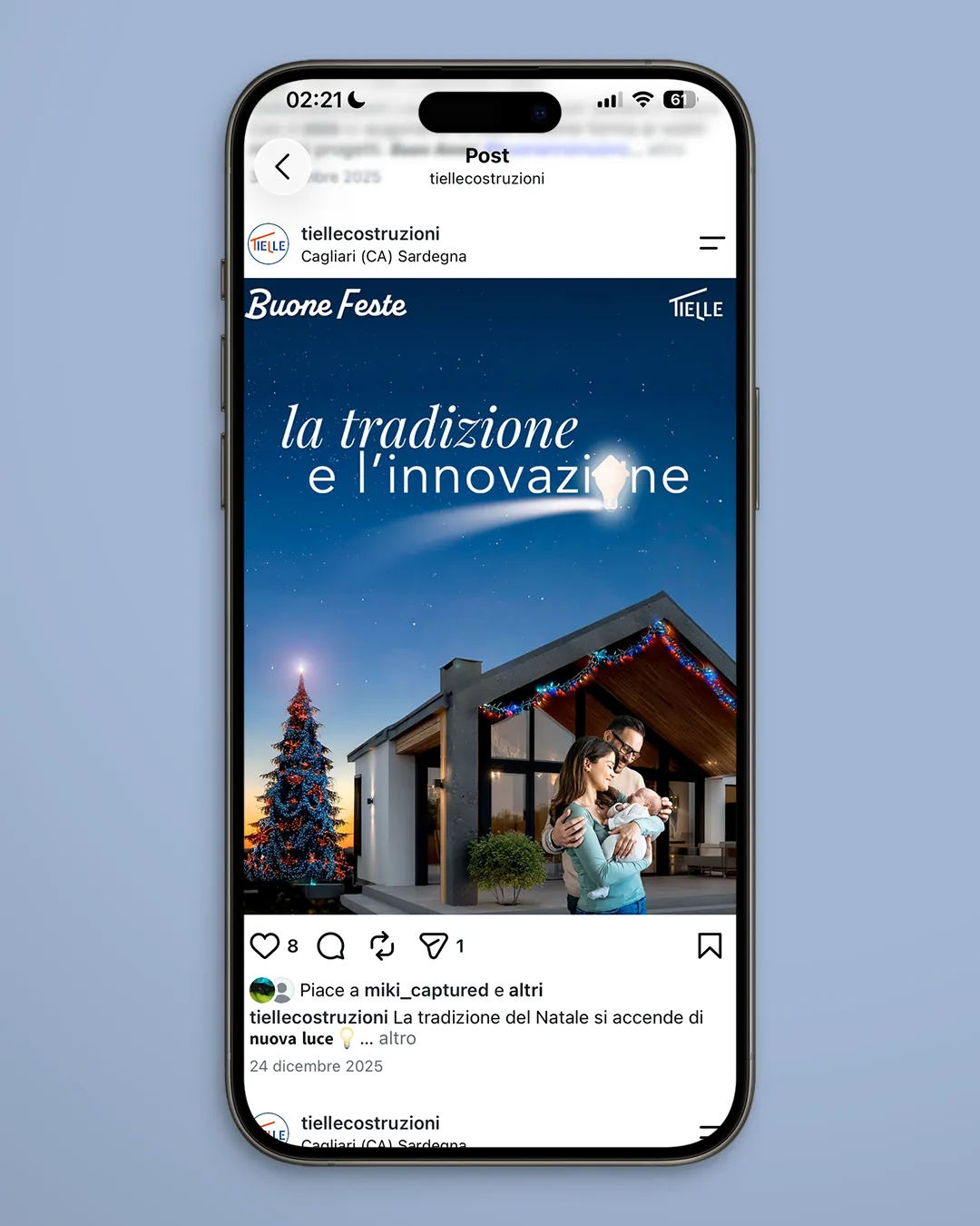 Écran d’Instagram sur un smartphone avec la publication de vœux de Tielle Costruzioni : l’inscription la tradition et l’innovation, un sapin de Noël surmonté d’une ampoule et un couple avec un nourrisson devant une maison ornée d’une guirlande de lumières