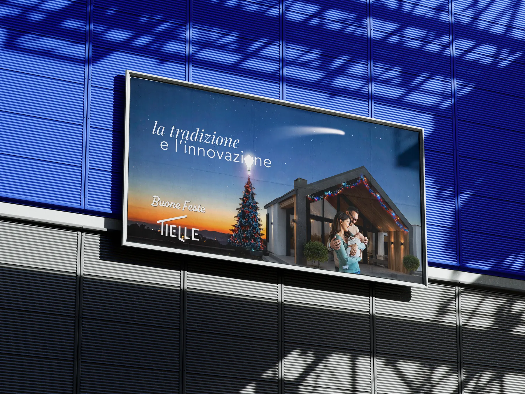Panneau sur une façade bleue avec le visuel de vœux : une famille avec un nourrisson devant une maison, un sapin de Noël surmonté d’une ampoule et l’inscription la tradition et l’innovation